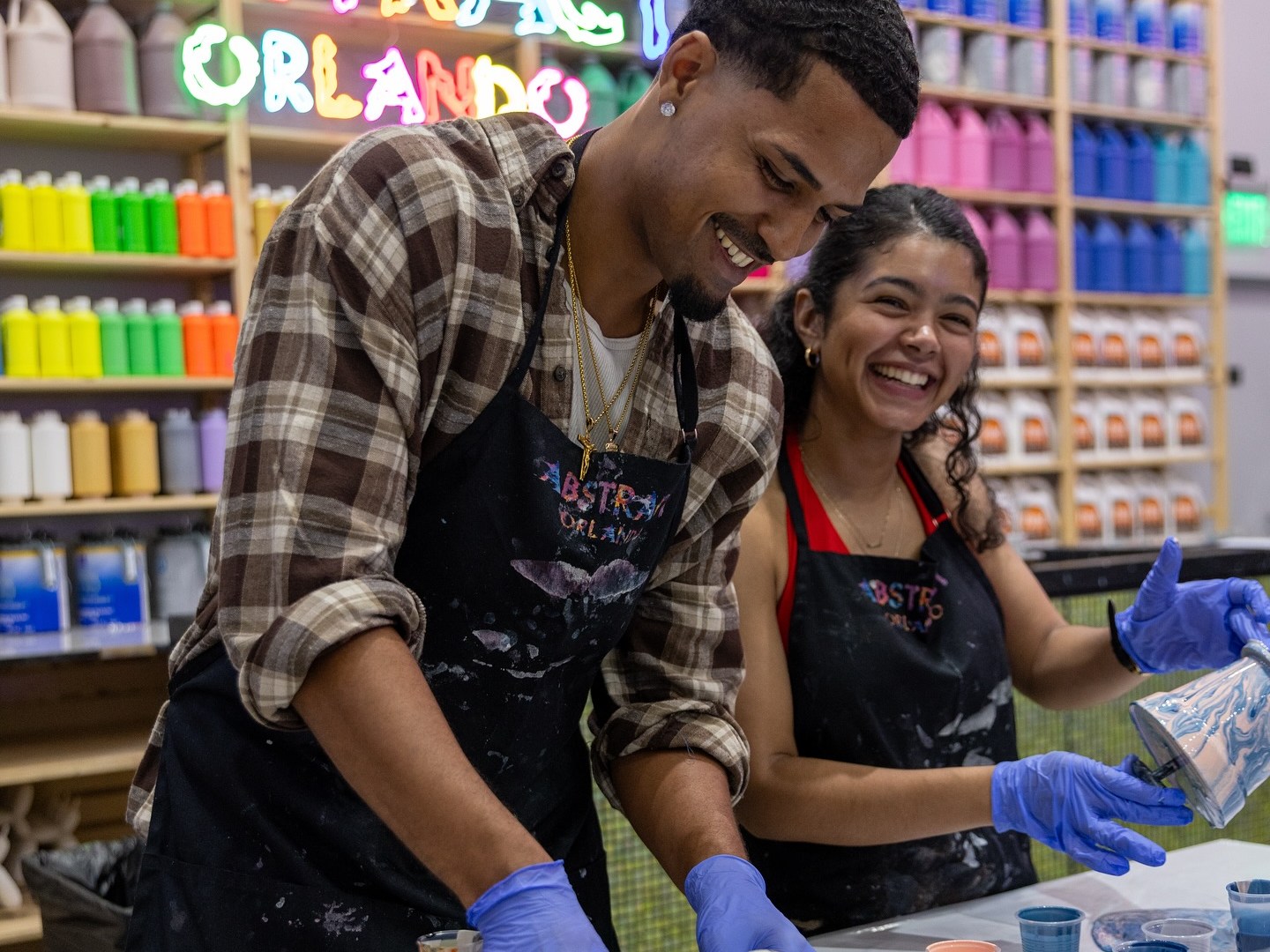 Two people in gloves enjoying painting, with colorful paints and a neon Abstract Orlando sign behind them.