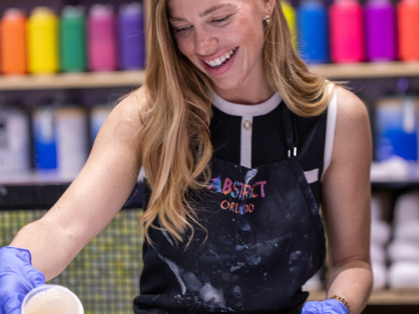 Woman smiling, wearing gloves, pouring liquid in art studio with colorful paint bottles on shelves.