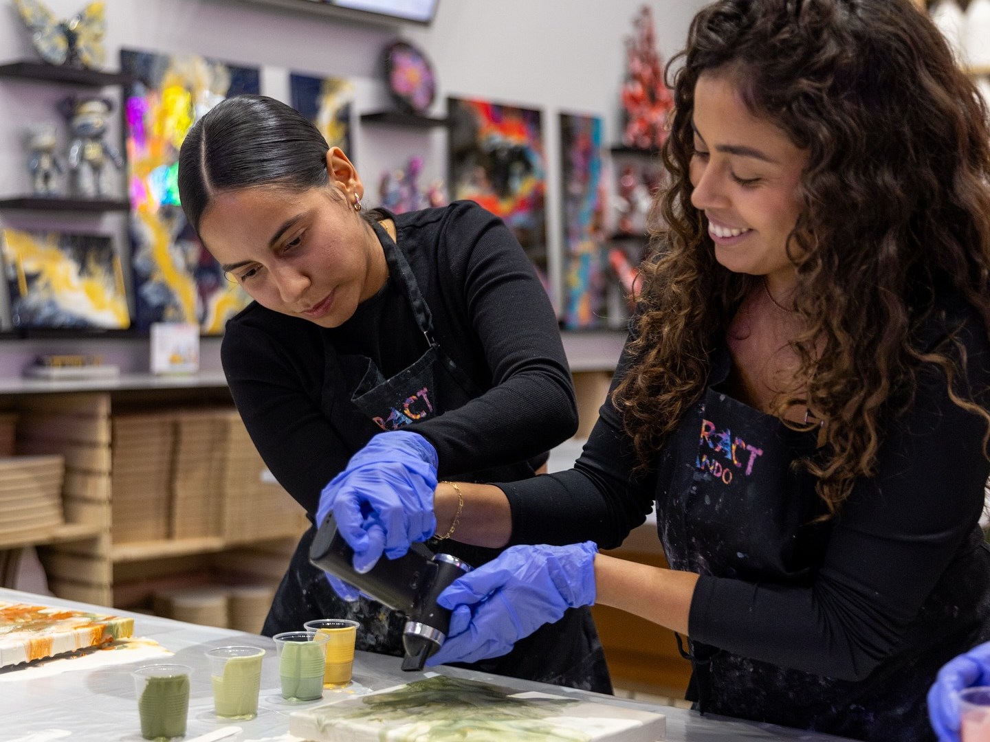 Two women in aprons and gloves painting together at a table covered with art supplies.