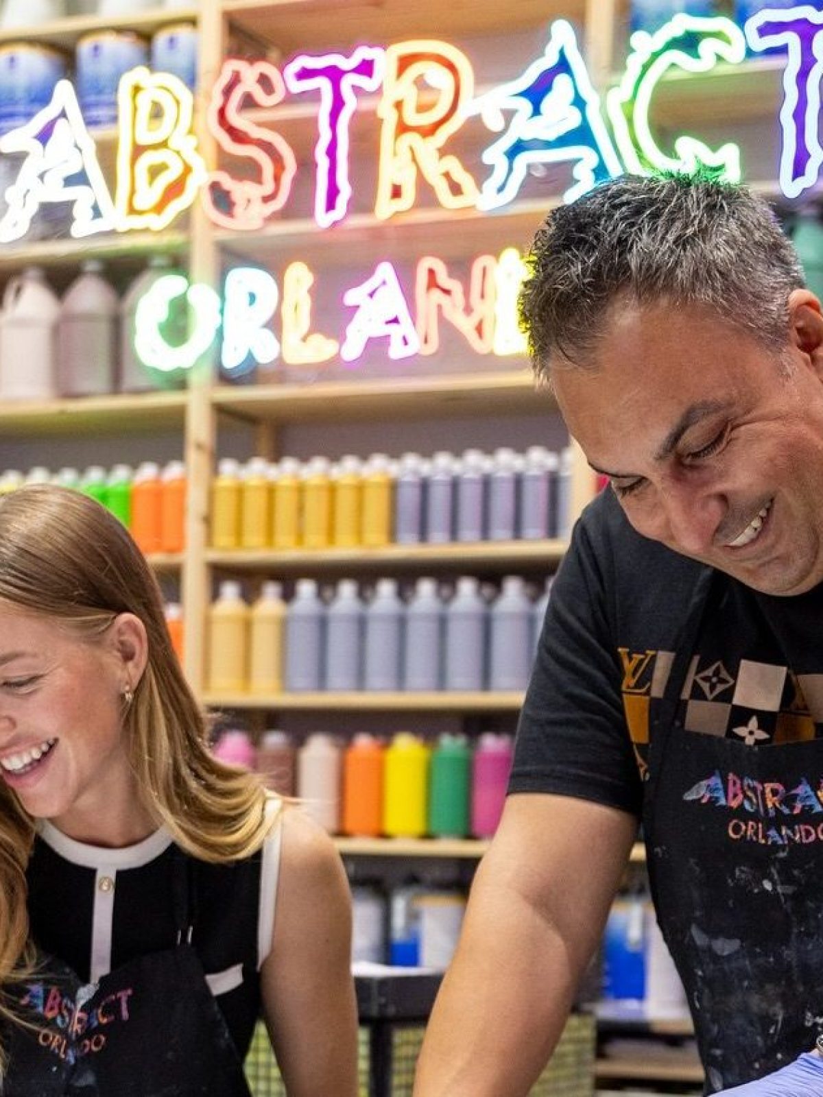 Two people pouring paint on canvases in art studio with colorful paint bottles and neon 'Abstract Orlando' sign.