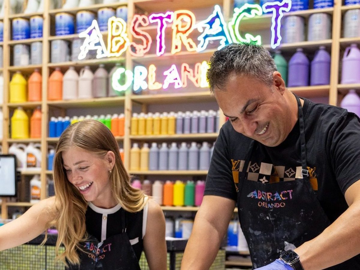 Two people pouring paint on canvases in art studio with colorful paint bottles and neon 'Abstract Orlando' sign.