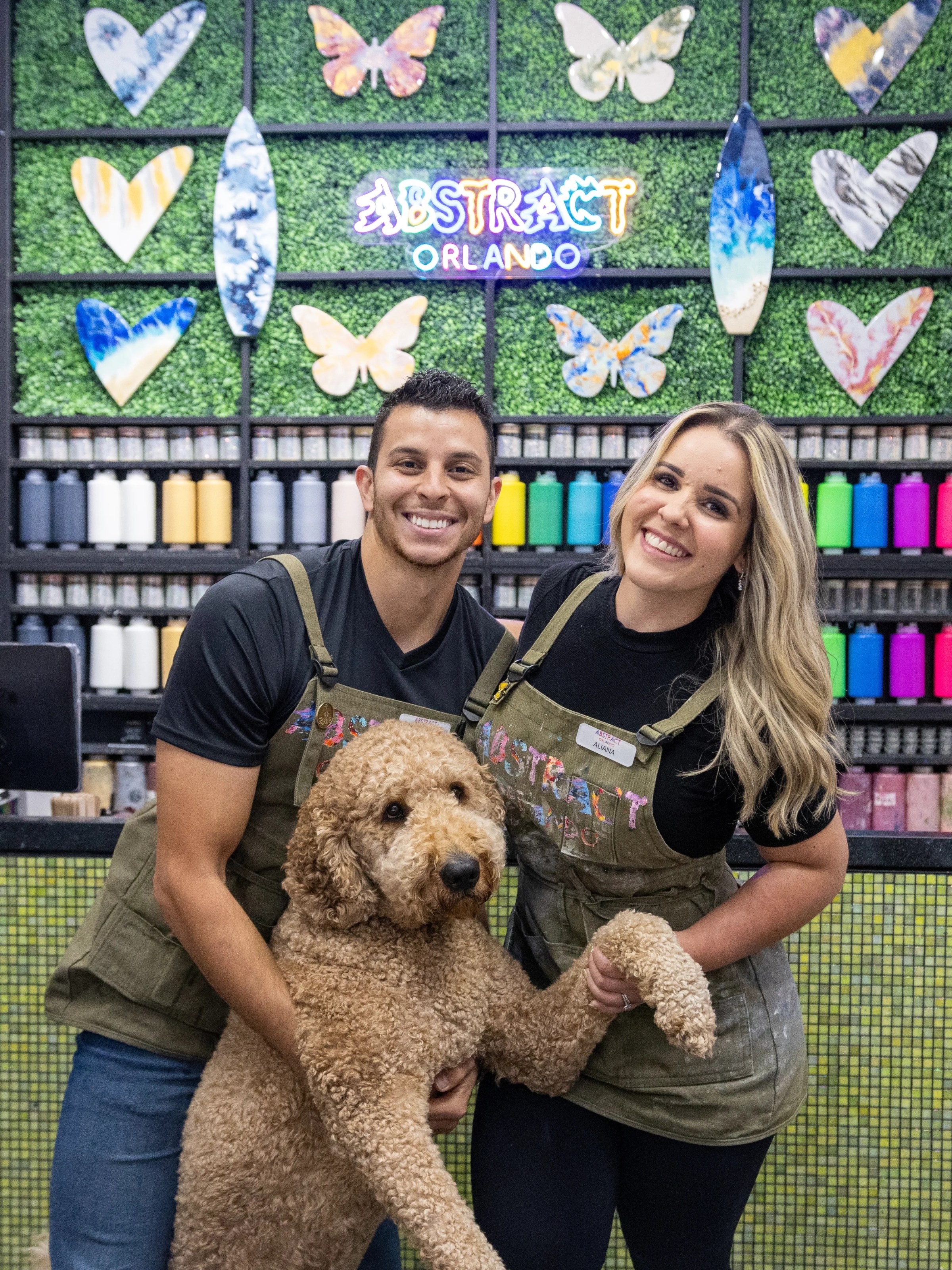 Two people and a dog smiling in front of colorful paints and decorations.