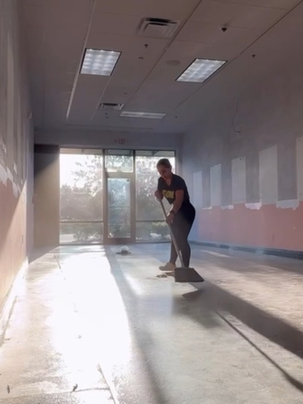 Person sweeping an empty, sunlit room with partially painted walls and bare floor.