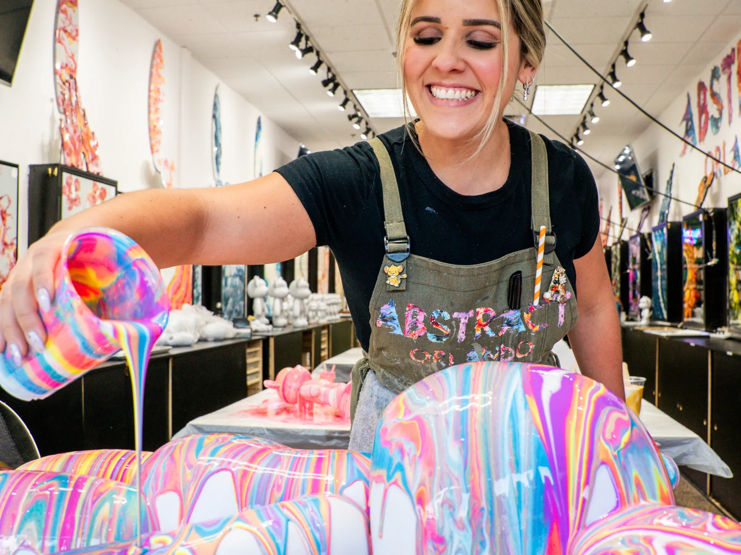 Person pouring vibrant multicolored paint over a 3D object, creating an abstract design. Smiling in an art studio.