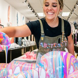 Person pouring vibrant multicolored paint over a 3D object, creating an abstract design. Smiling in an art studio.