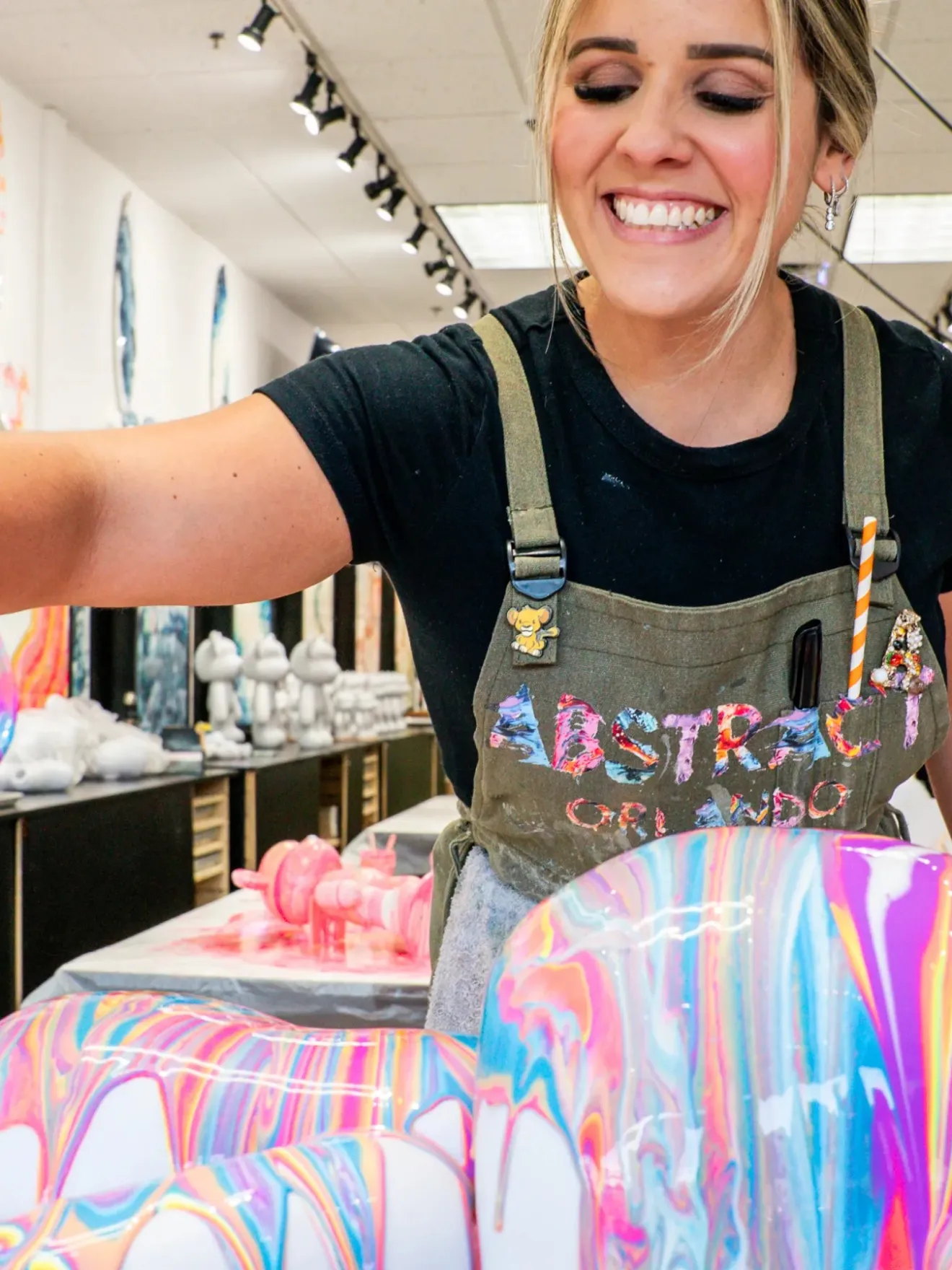 Smiling woman pours colorful paint over abstract sculptures in an art studio.
