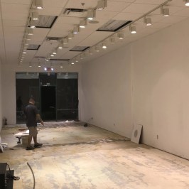 Person in an empty room with exposed flooring and overhead lights.