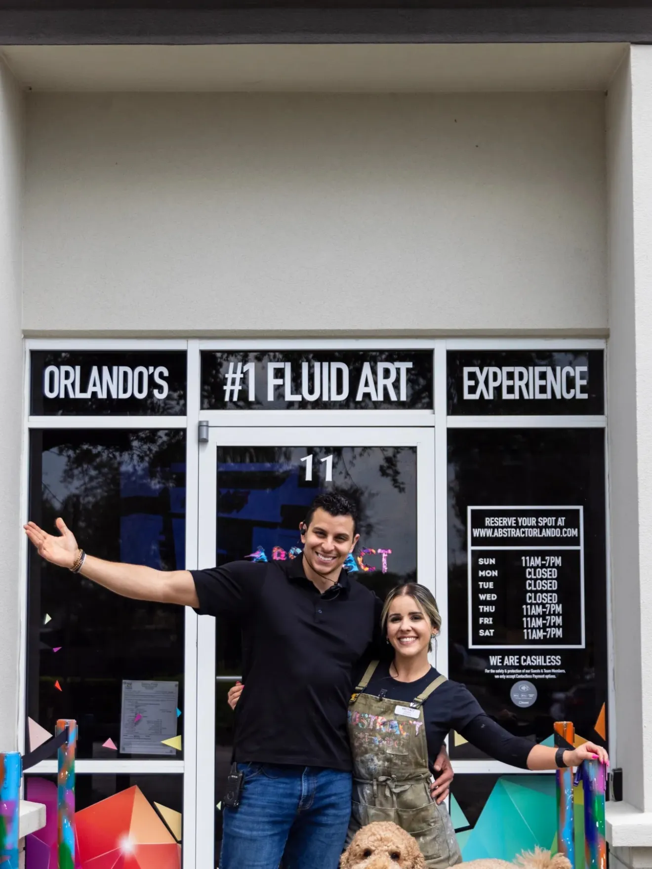 Two people stand smiling in front of a fluid art studio entrance with colorful decor.
