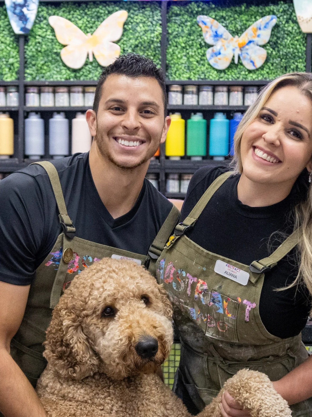 Two people smiling with a dog in front of colorful paint bottles and decorated wall.