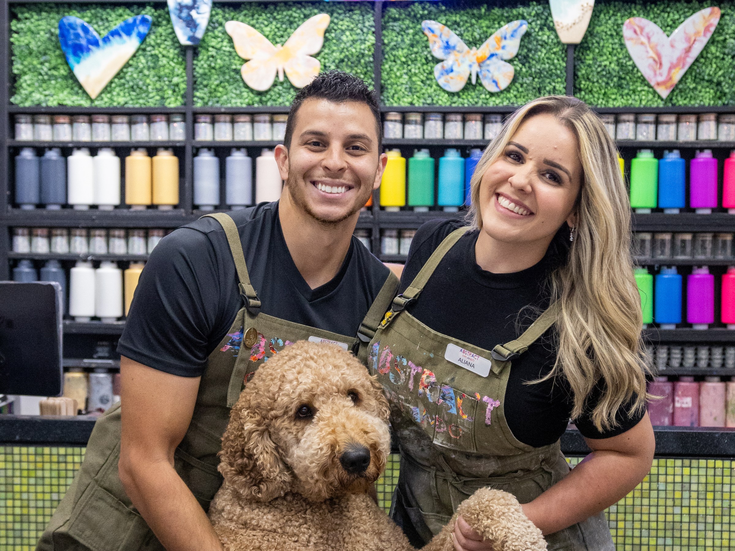 Two people smiling with a brown curly-haired dog in front of a colorful wall with art supplies and decorations.