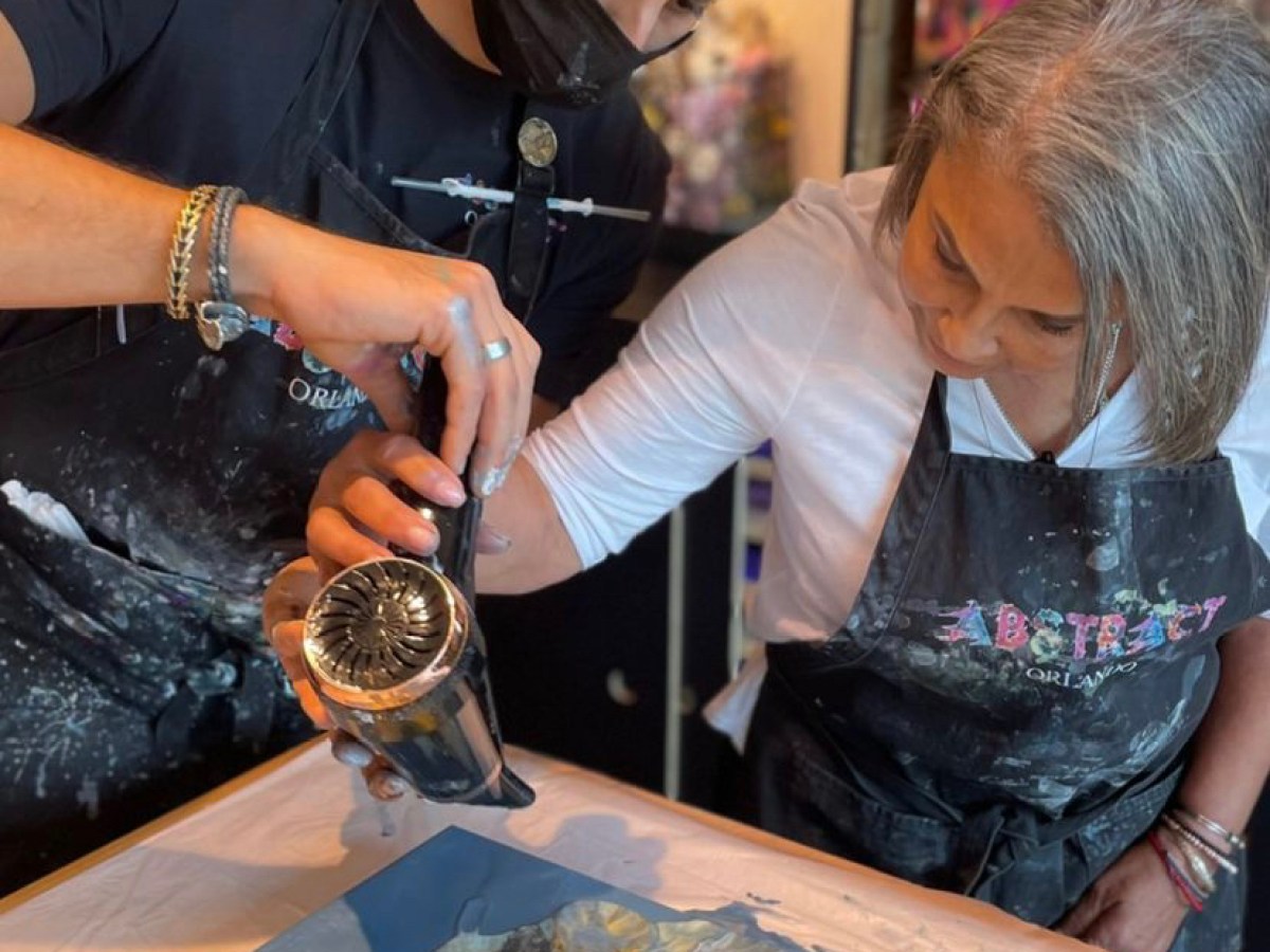 Two people using a hairdryer to spread paint on a canvas.