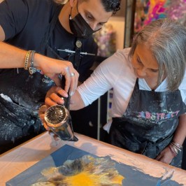 Two people using a hairdryer to spread paint on a canvas.