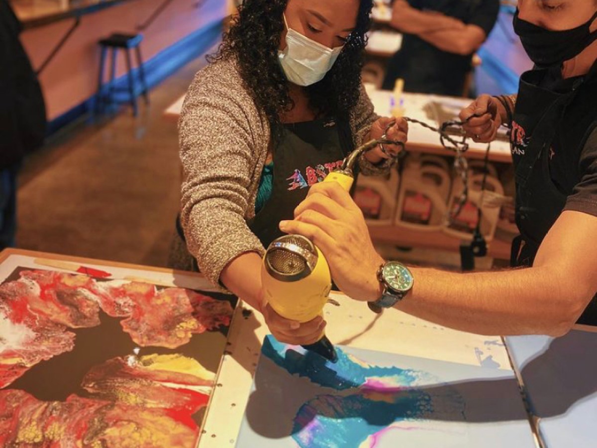 People using a heat tool on colorful artwork at an art workshop, both wearing masks.