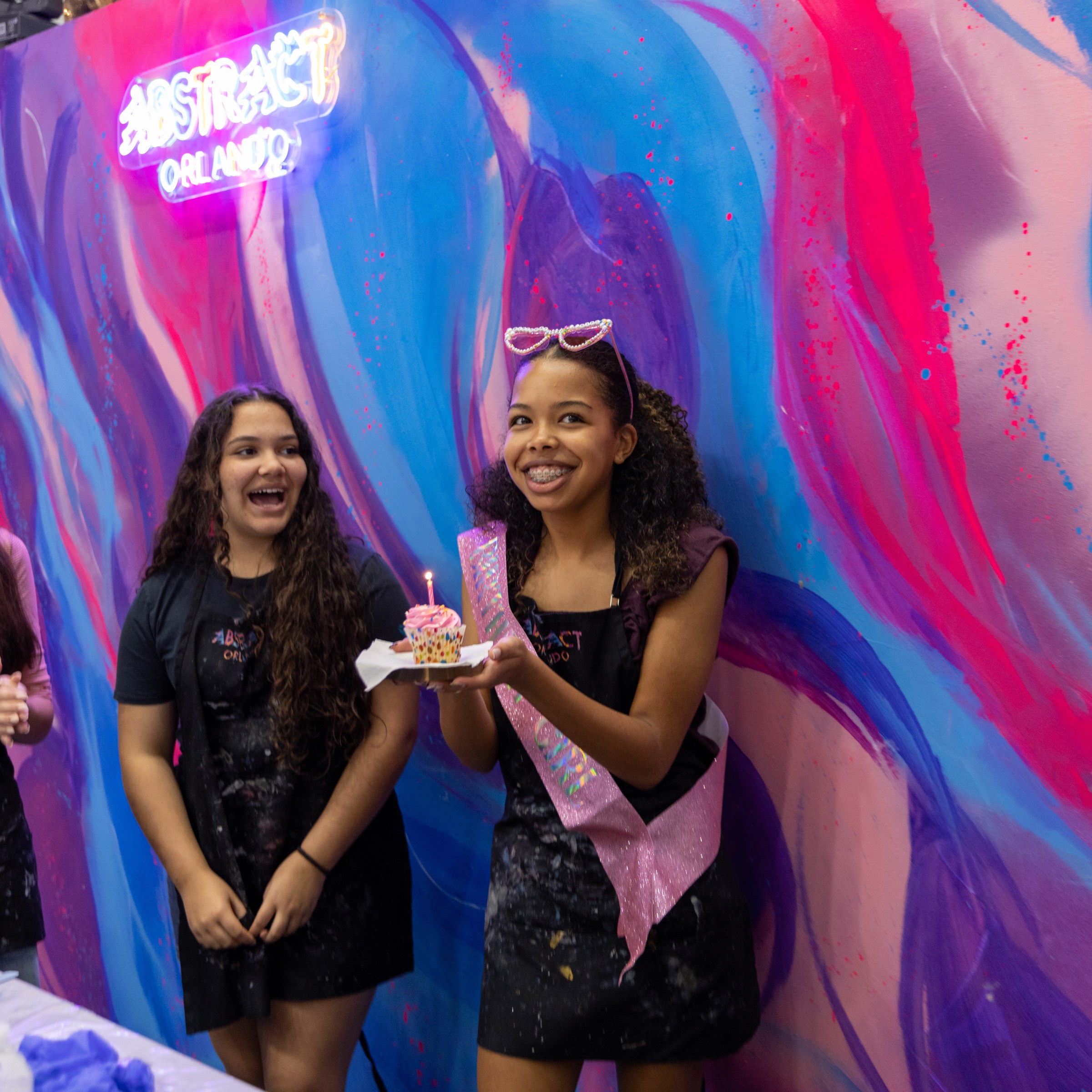 Two girls smiling, one holding a cupcake with a candle, in front of a colorful mural at Abstract Orlando.