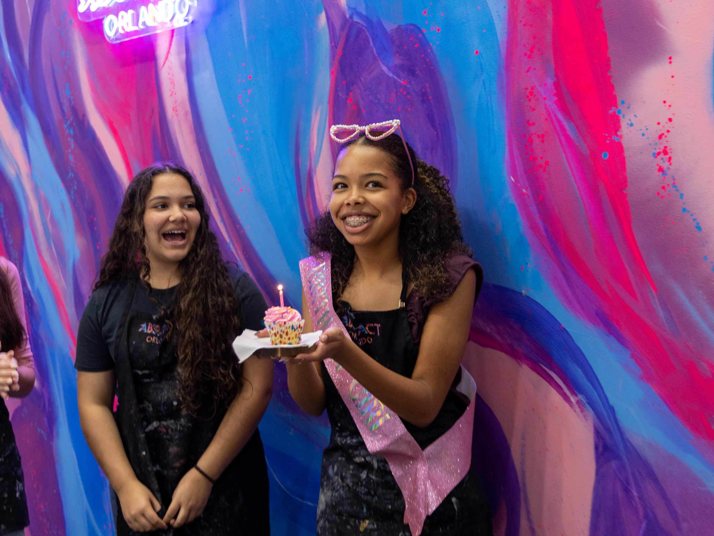 Two girls smiling, one holding a cupcake with a candle, in front of a colorful mural at Abstract Orlando.