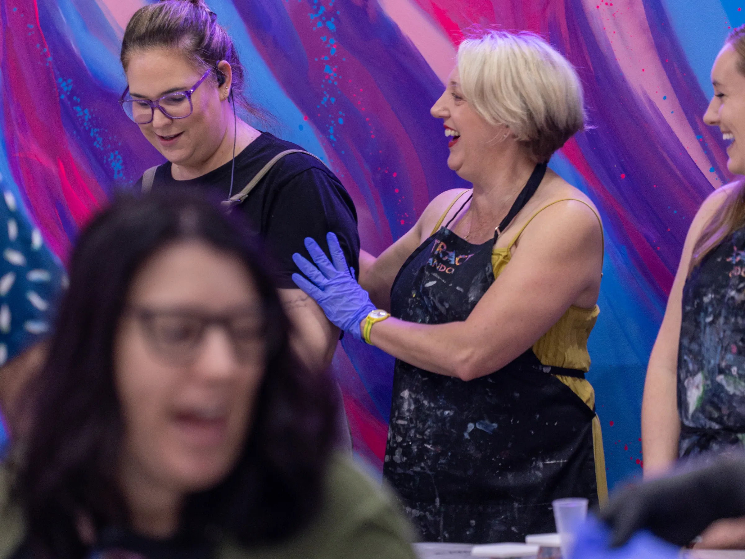 Women in aprons and gloves enjoy a painting workshop with vibrant abstract art on walls.