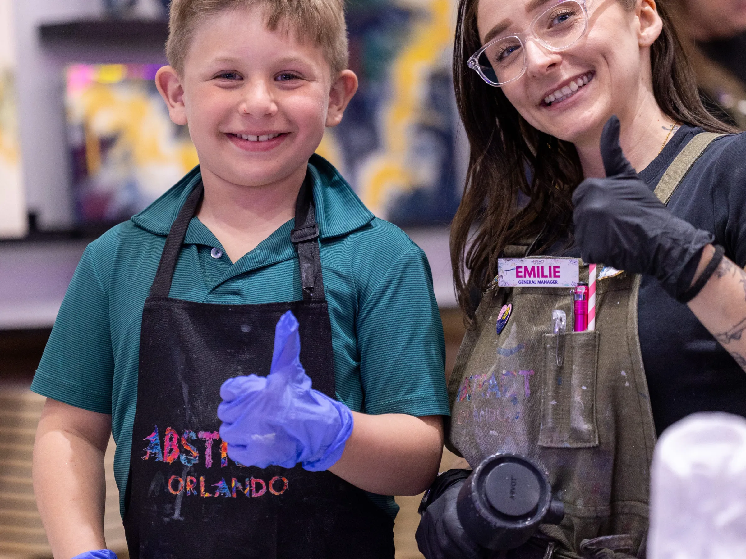Smiling child and adult with paint aprons giving thumb up in art studio.