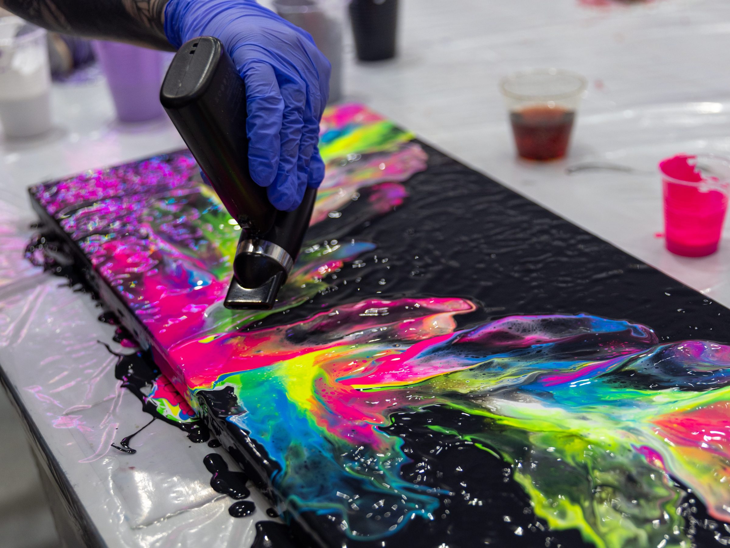 Person applying colorful paint to canvas with a plastic tool, on a table covered with plastic.