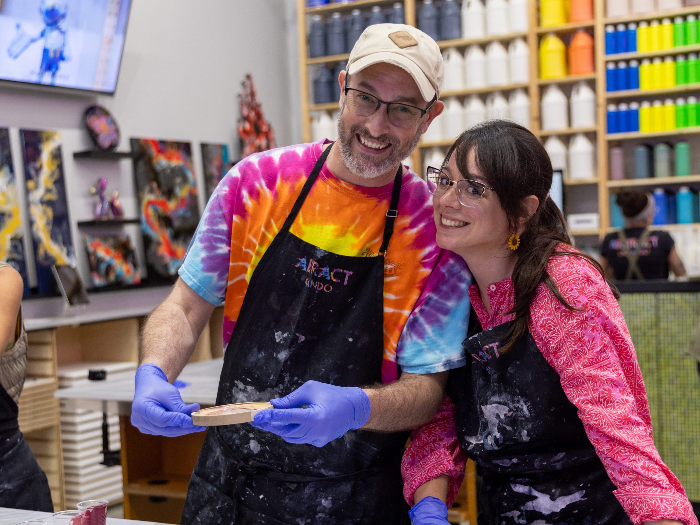 Two people in aprons smiling, standing by a table with paint supplies.