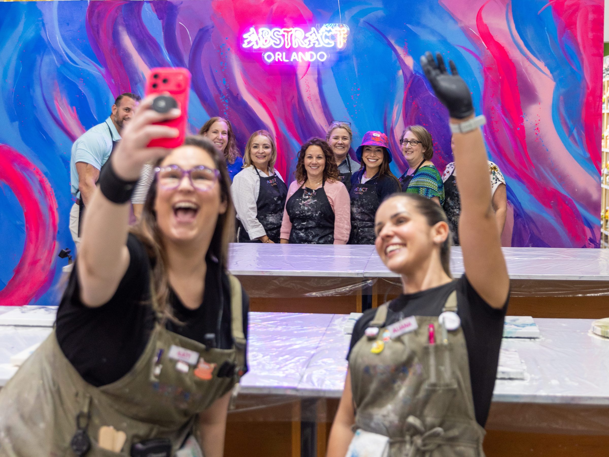Group takes selfie at colorful art venue with neon sign and painted backdrop.