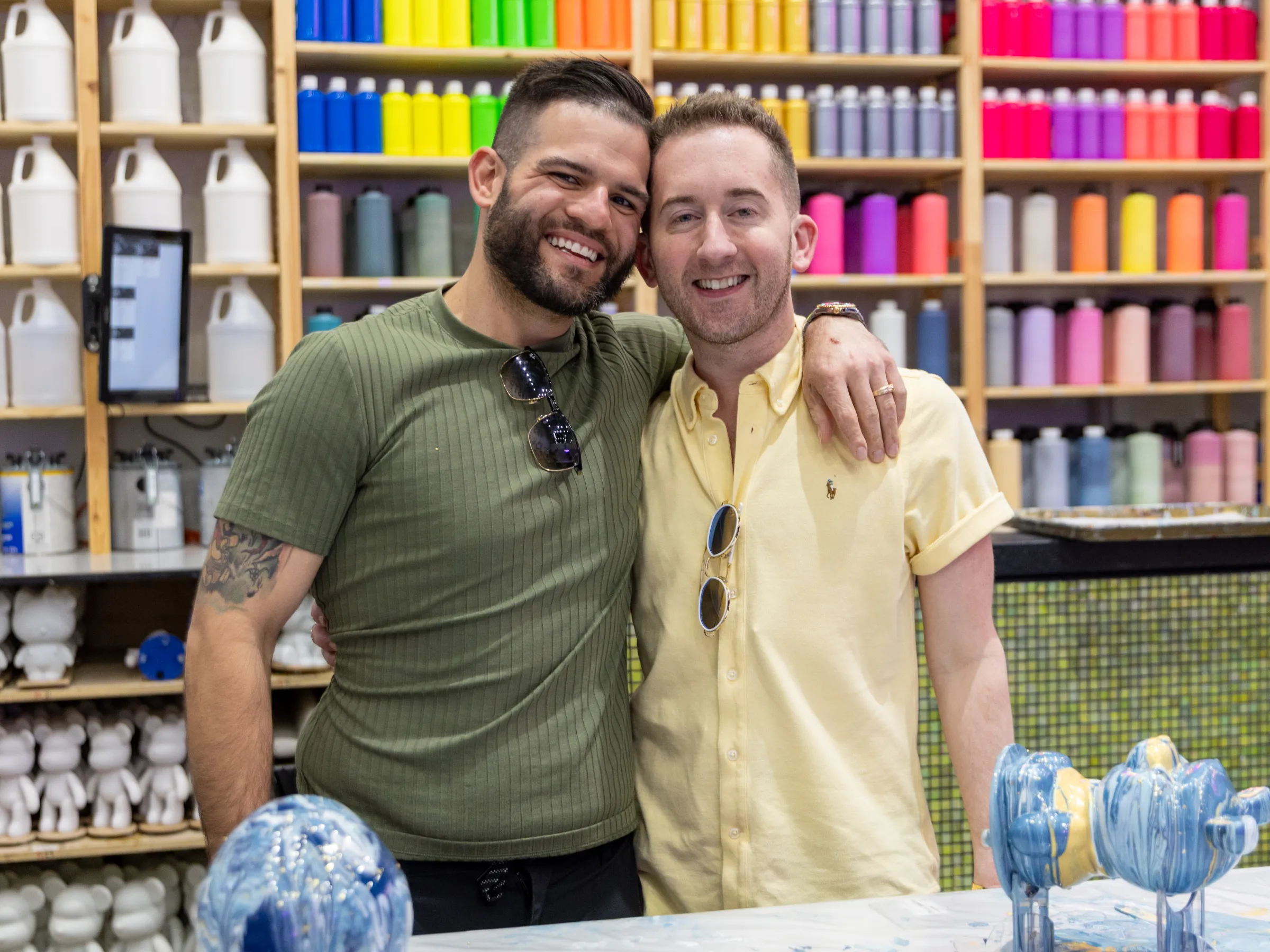 Two men smiling in art studio with colorful paint bottles on shelves.