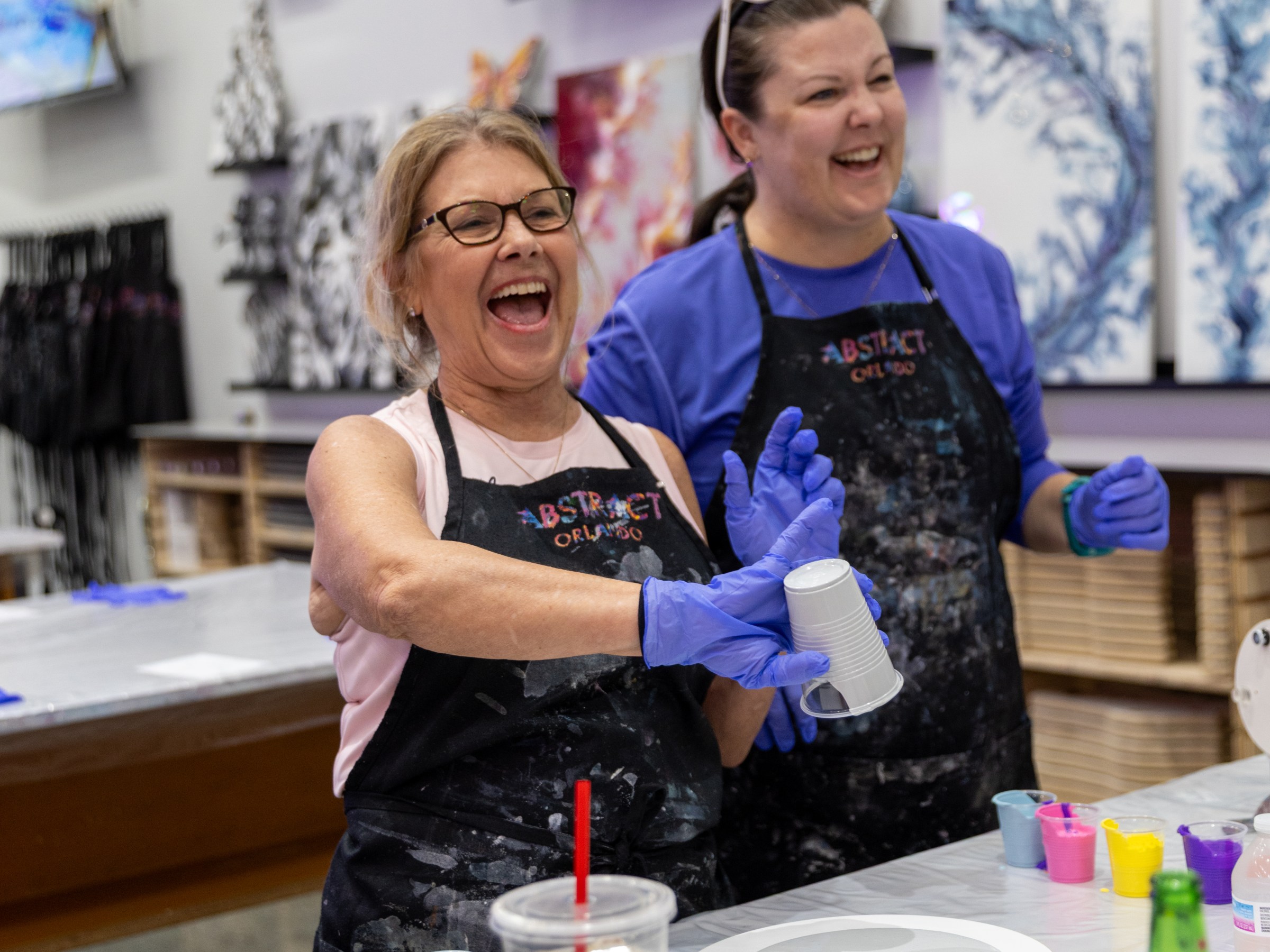 Two women laughing, wearing aprons, at an art studio with paint supplies.