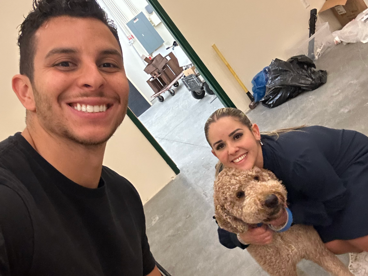 Two people smiling with a curly-haired dog indoors.