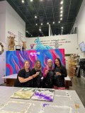 Four women holding drinks in an art studio with colorful paintings and decorations.