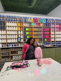 Two people smiling in an art studio with colorful paint bottles on shelves.