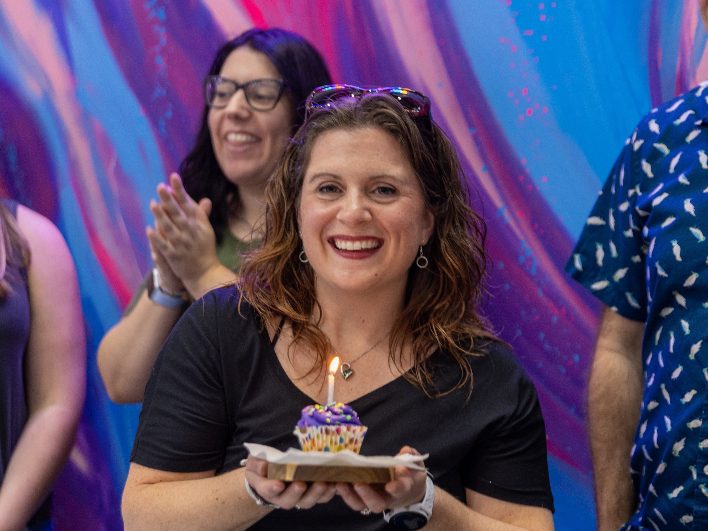Woman smiling, holding a cupcake at a colorful event.