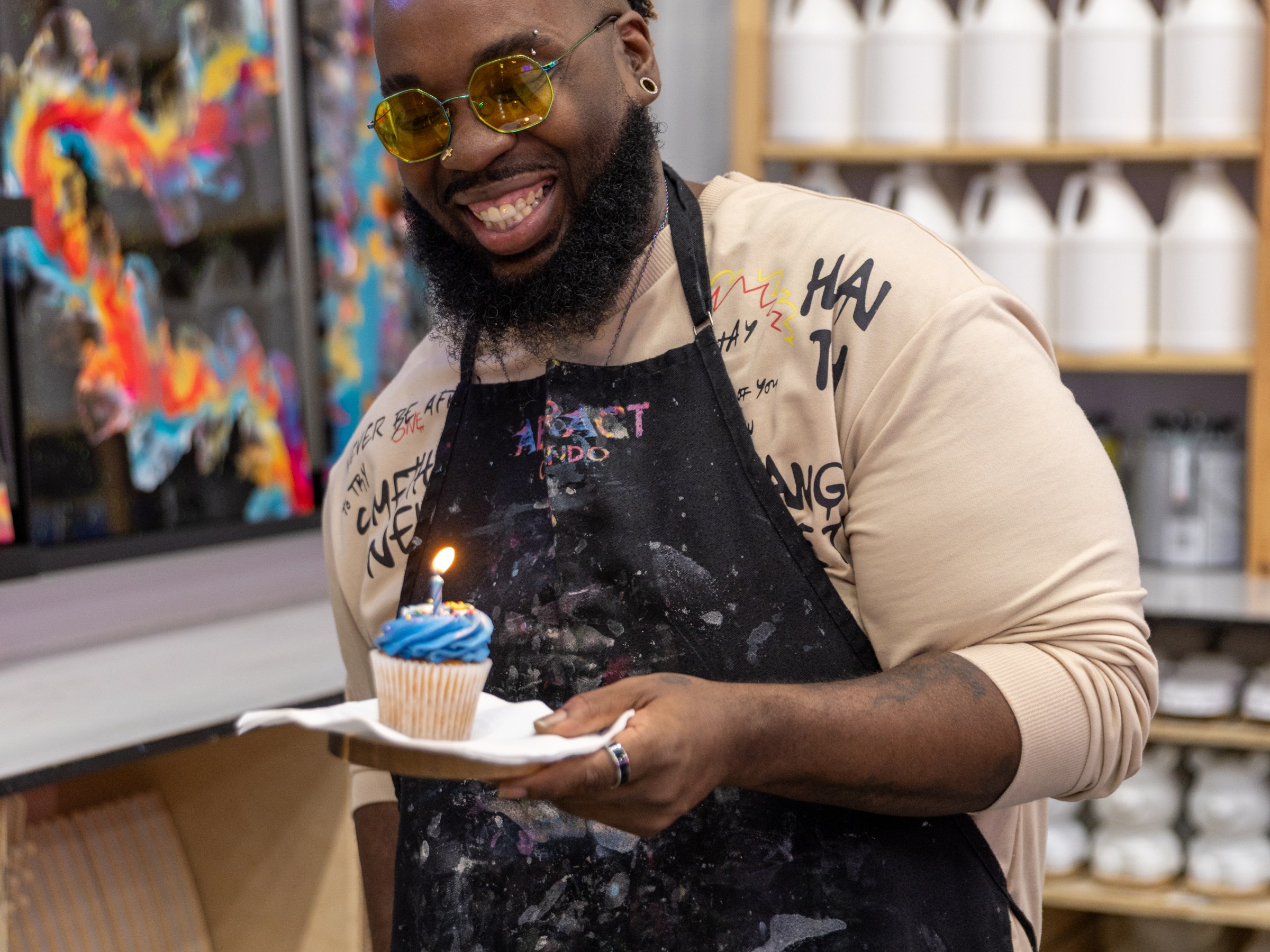 Smiling person holding a cupcake with a lit candle in an art studio.