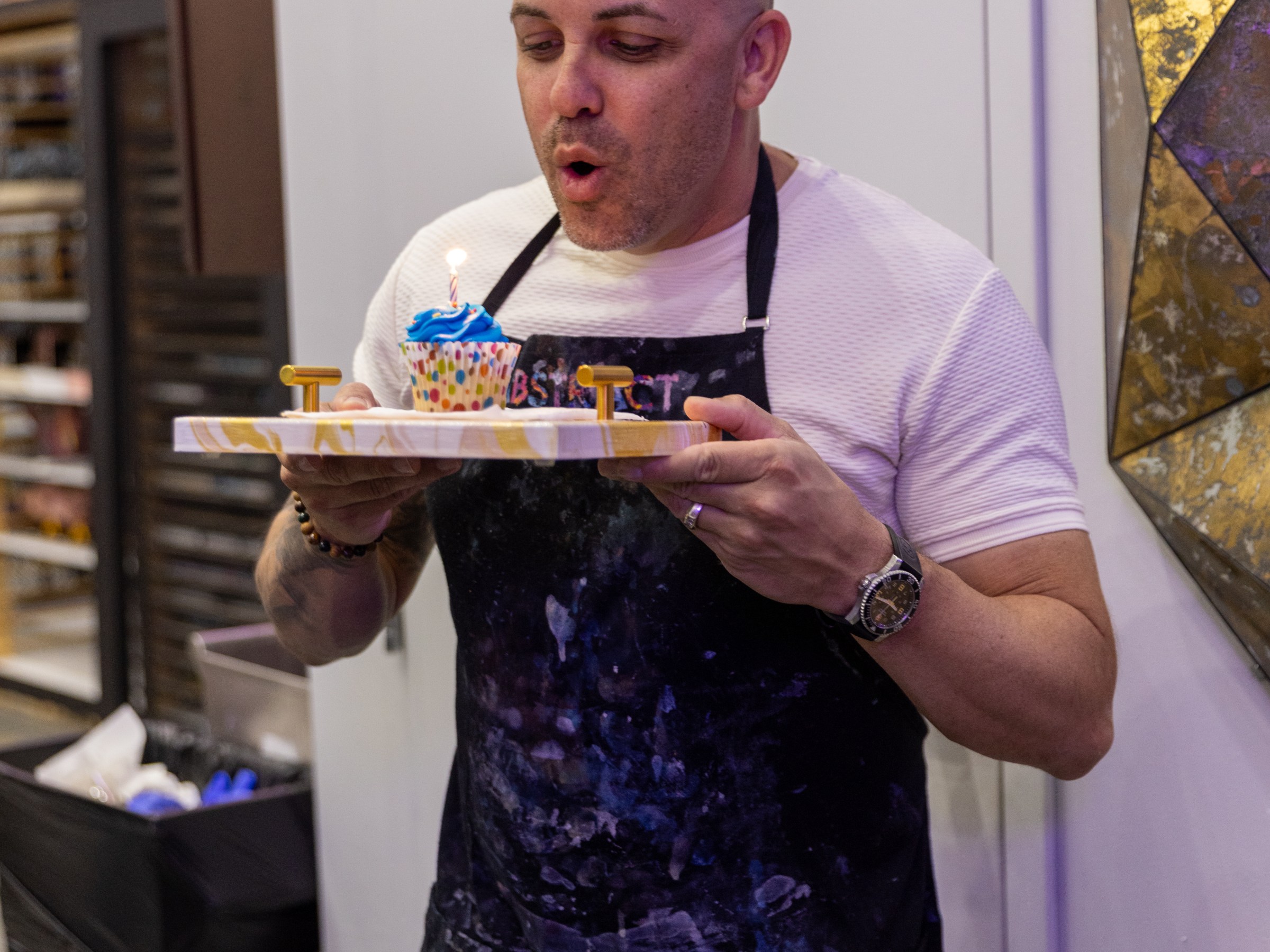 Man in apron blowing candle on a cupcake.